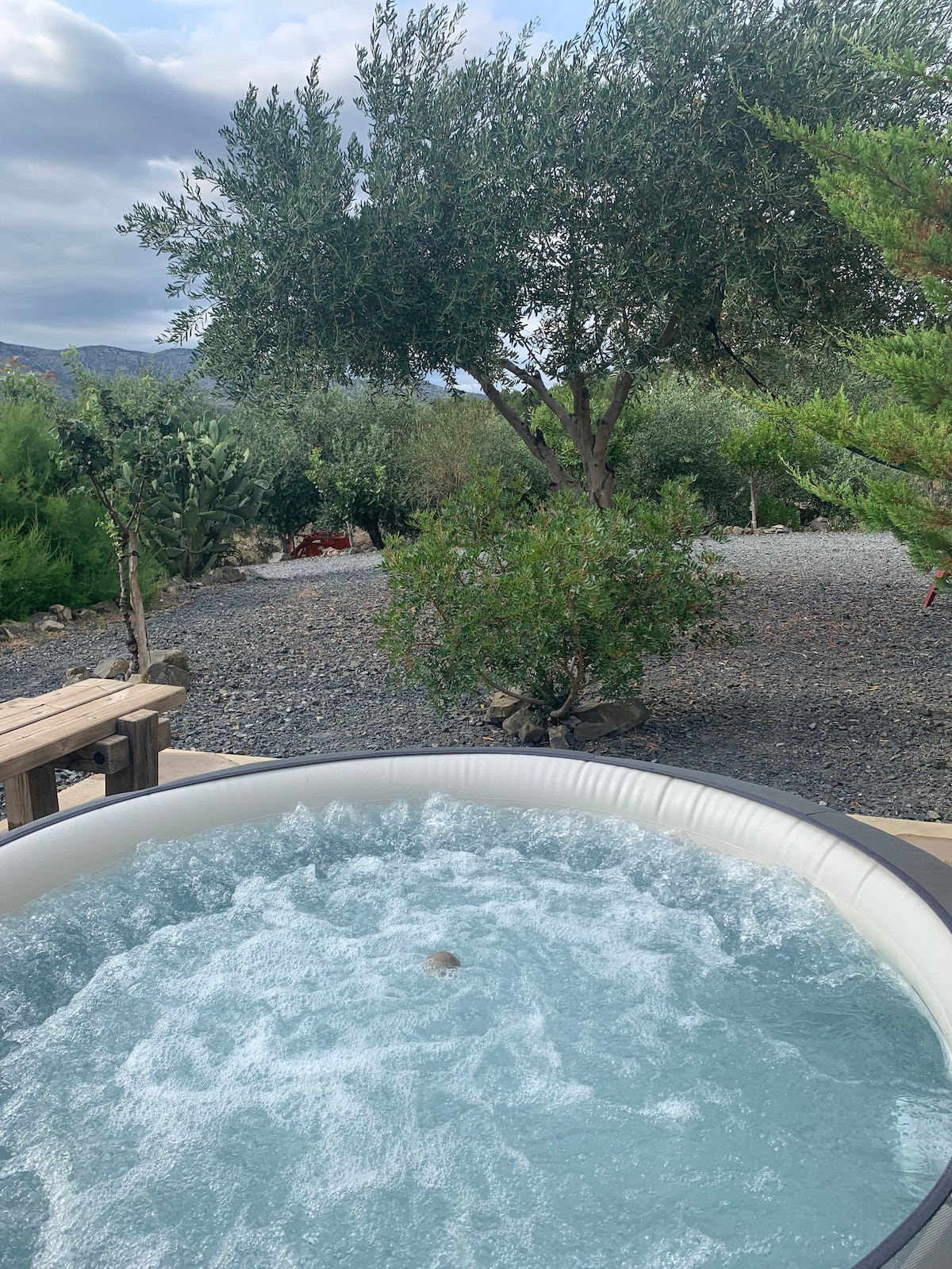 A private hot tub is shown, surrounded by lush greenery and olive trees. The bubbling water creates a serene atmosphere, with natural stones and plants visible in the background. A wooden deck appears adjacent to the hot tub, offering an inviting space for relaxation.