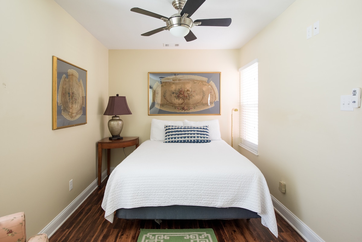 A well-lit bedroom features a spacious bed adorned with a white coverlet. Two framed artworks hang above the bed, complemented by a bedside table with a lamp. A ceiling fan is present, and natural light enters through a partially covered window.