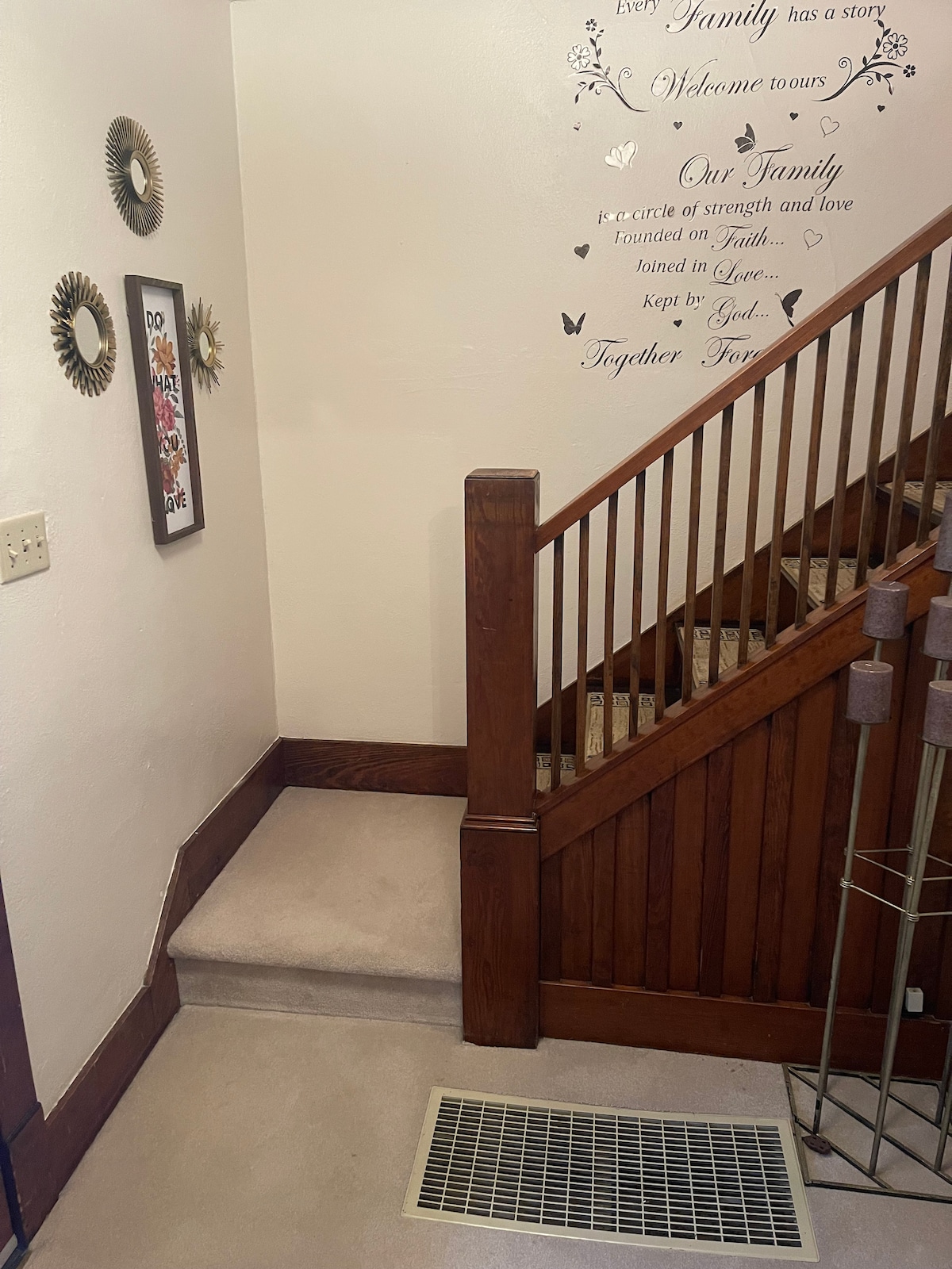 The entrance features a wooden staircase with a detailed handrail, leading to an upper level. A framed wall art piece is displayed beside the stairs, and decorative mirrors are arranged on the adjacent wall. Soft carpeting covers the steps for added comfort.