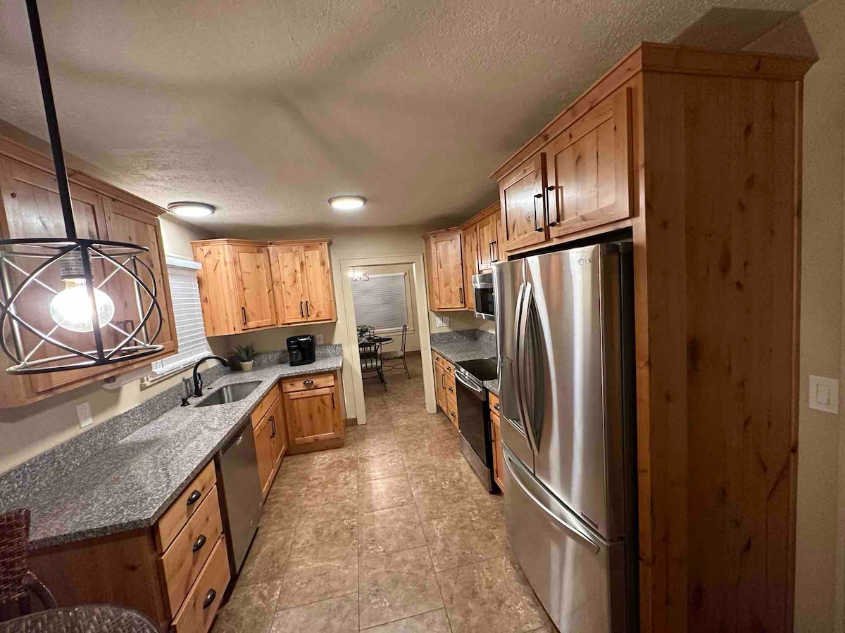 A well-equipped kitchen features wooden cabinetry and granite countertops, along with stainless steel appliances. A sink is positioned beneath a window, and a coffee maker is visible. Natural light illuminates the room from overhead fixtures, enhancing the inviting space.