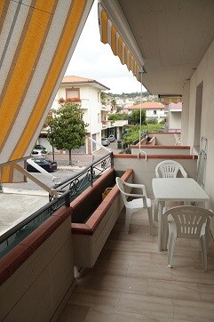 A spacious balcony is presented, featuring a white dining table with matching chairs and a decorative awning providing shade. A view of the surrounding neighborhood is visible, showcasing nearby buildings and street activity. A potted plant adds a touch of greenery to the space.