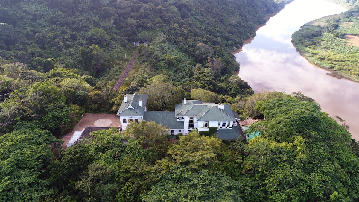 An aerial view captures the Royston Hall Guest House nestled in lush greenery, surrounded by trees and overlooking a winding river. The property features a spacious structure with a green roof, blending harmoniously with the natural landscape.
