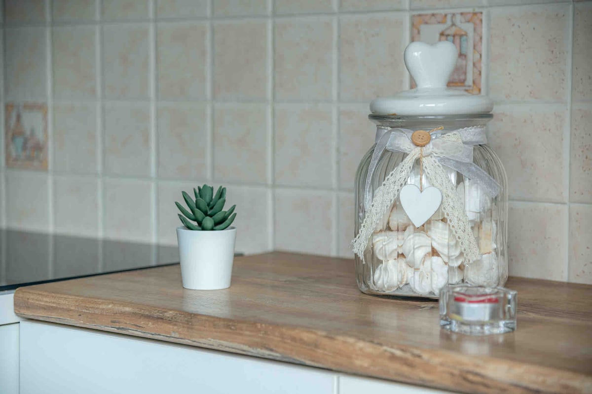 A kitchen countertop is depicted featuring a glass jar filled with meringues, adorned with a decorative ribbon and a heart tag. A small potted succulent is placed nearby, complementing a simple candle holder. The background showcases light-colored tiled walls.
