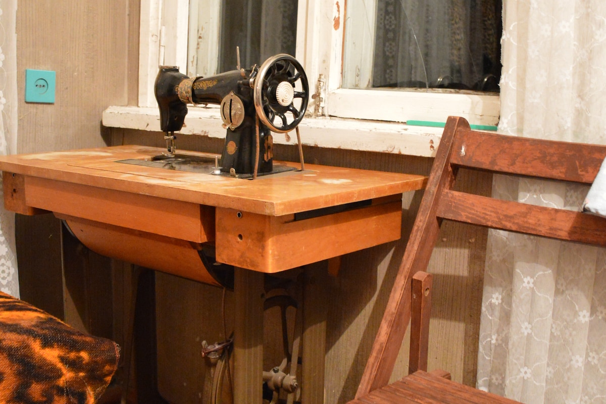 An antique sewing machine is positioned on a wooden table, complemented by a rustic wooden chair. The scene is illuminated by light from a nearby window, with sheer curtains providing a soft, diffused effect.