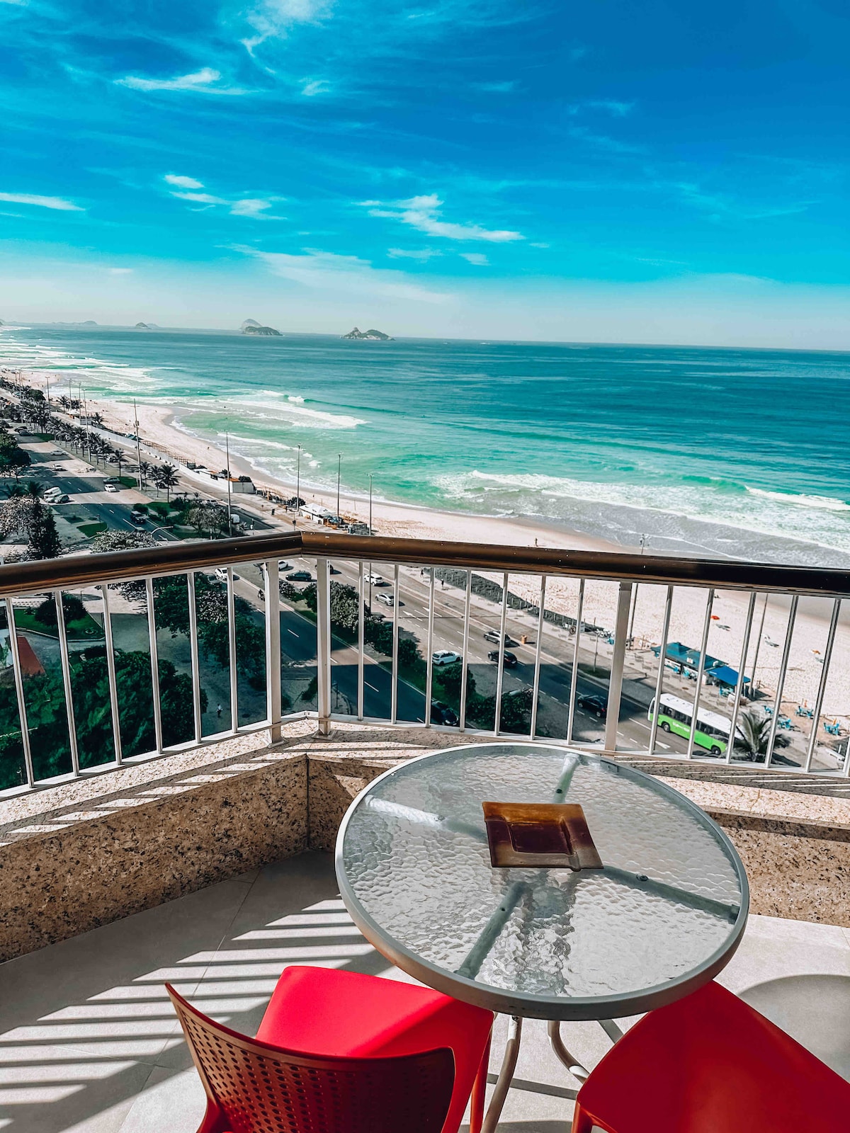 A balcony view features a round glass-top table with two red chairs, overlooking the expansive sandy beach and vibrant blue ocean. Soft waves are seen lapping the shore, with distant mountains framed against the clear sky.