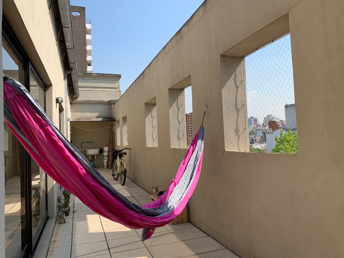 A spacious terrace is presented with a colorful hammock draped between two walls. The flooring is made of light-colored tiles, and windows allow natural light to enter. A bicycle is positioned nearby, and greenery can be seen through the openings.