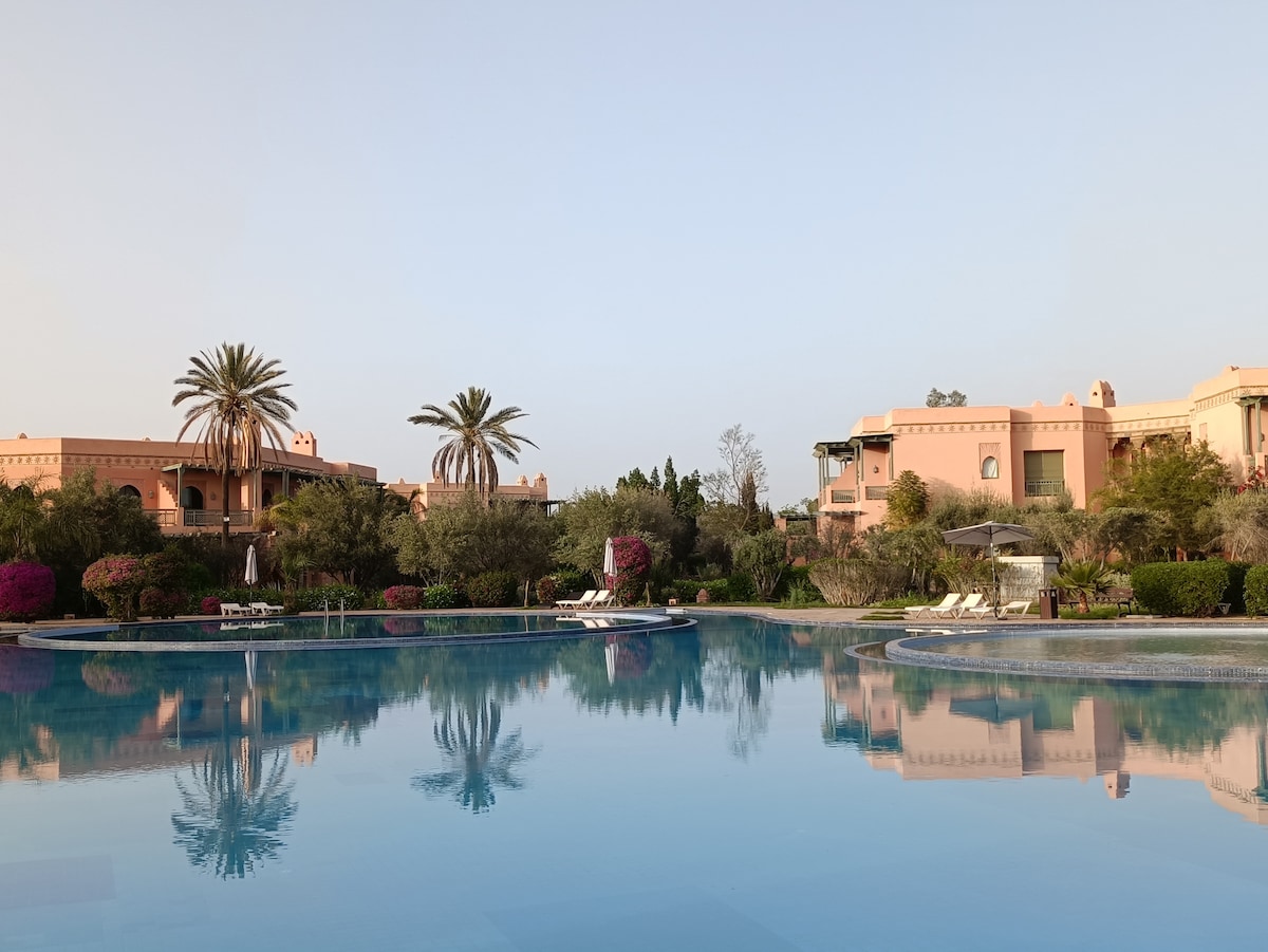 The serene pool reflects the surrounding architecture and vibrant greenery, framed by scattered palm trees and blooming flowers. Lounge chairs are positioned nearby, inviting relaxation under clear skies. The tranquil setting emphasizes the peaceful ambiance of the property.
