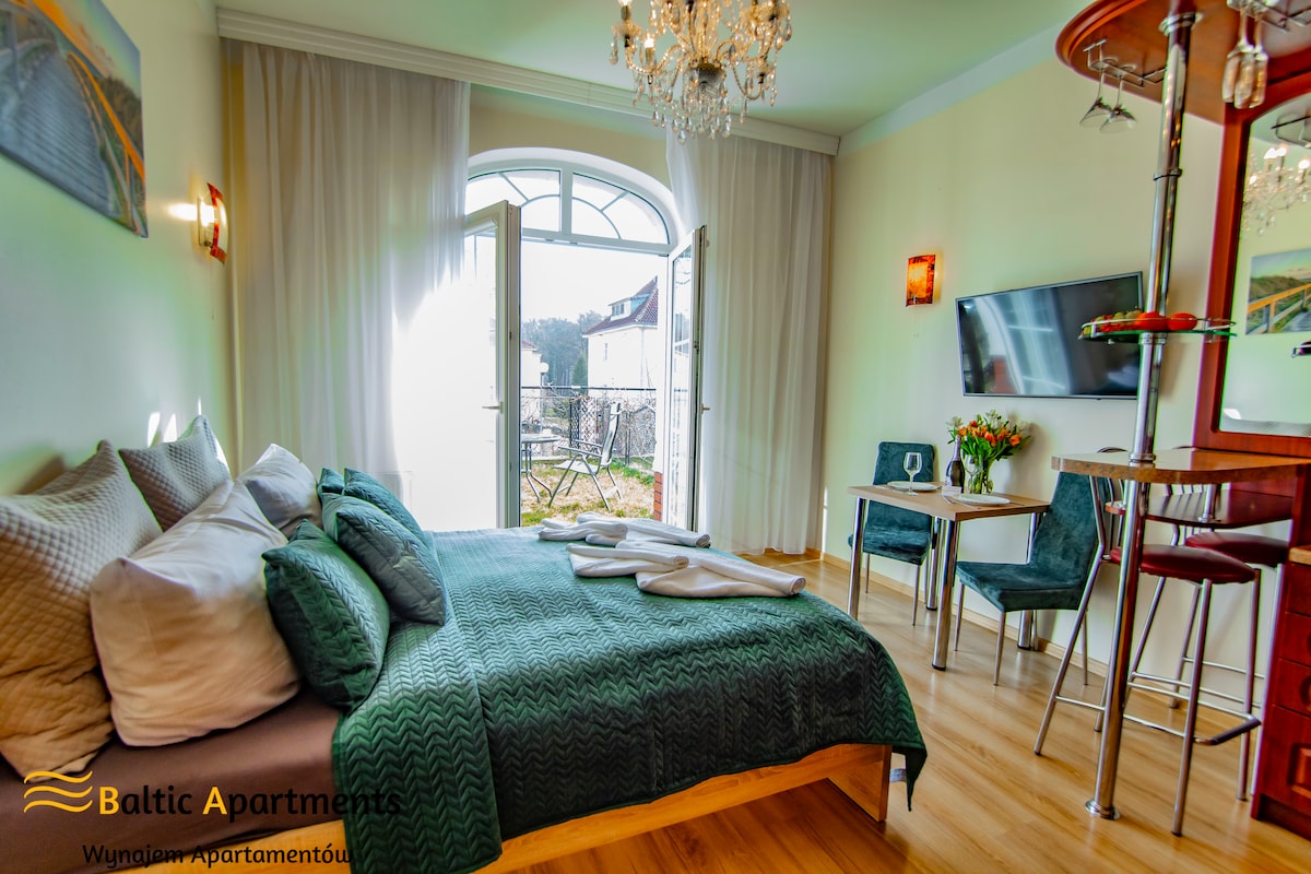 A spacious bedroom features a large double bed adorned with textured bedding. Natural light enters through an arched window, highlighting the modern décor. A small dining area with tall chairs is visible, along with a wall-mounted television and tasteful floral arrangements.