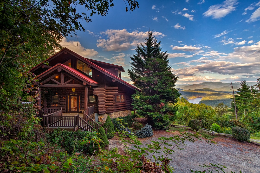 The exterior of the cabin is showcased against a backdrop of mountains and a lake. The structure features natural wood logs and a red roof, with a wide porch and steps leading to the entrance. Surrounding landscaping includes shrubbery and trees.