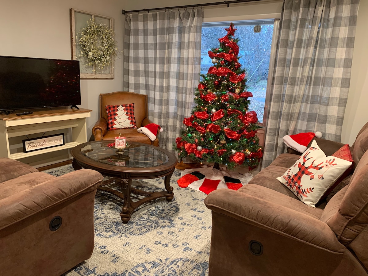A cozy living area features two recliner couches positioned around a glass coffee table. A television is placed on a light-colored console. A decorated Christmas tree stands near a window, accompanied by seasonal decorations on the floor.