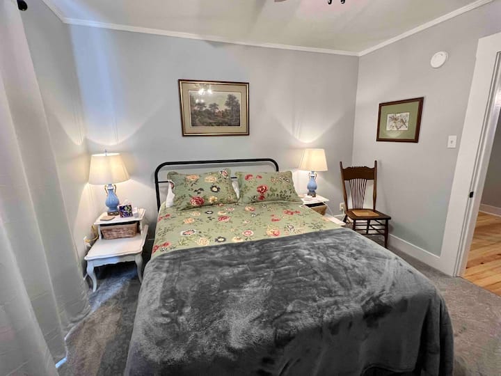 The master bedroom has a queen bed, wardrobe for clothes, ceiling fan, wall-mounted TV, end tables, charging blocks, chair, luggage rack, lamps and direct access to a full bath.