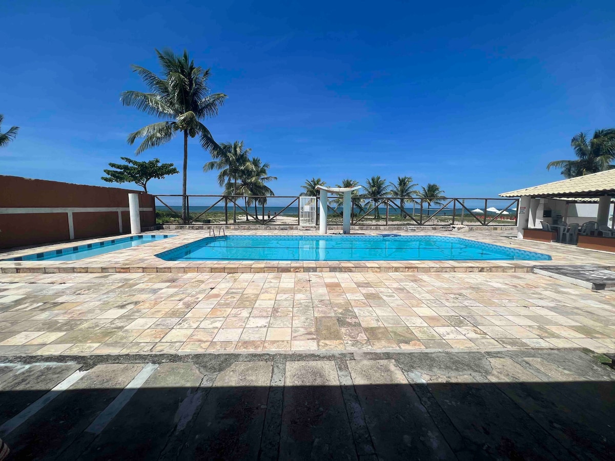 A large outdoor pool is surrounded by a spacious deck, adorned with palm trees in the background. A covered seating area is visible to the right, providing shade. The clear blue sky and calm ocean enhance the relaxation offered by this inviting space.