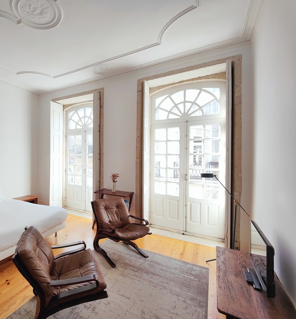 Natural light fills the room through two tall, arched windows, enhancing the soft tones of the walls. Two brown leather chairs are positioned beside a wooden desk, creating a cozy seating area. A minimalist decor style is evident, with wooden flooring complementing the space.