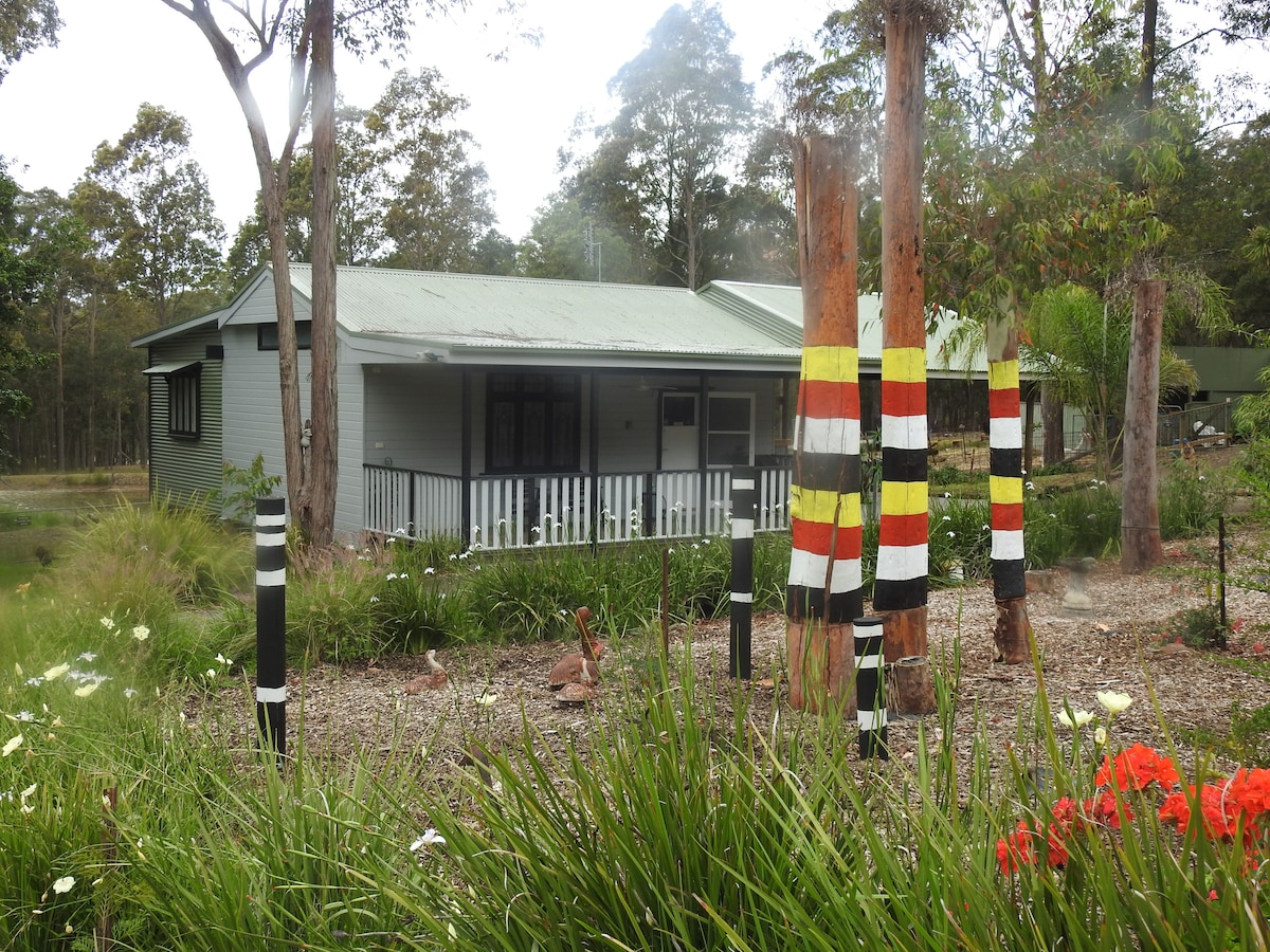 A cottage is surrounded by lush greenery and colorful plant beds. Indigenous art adorns trees in the foreground, featuring bold stripes of black, red, and yellow. The building has a welcoming porch and large windows that invite natural light.