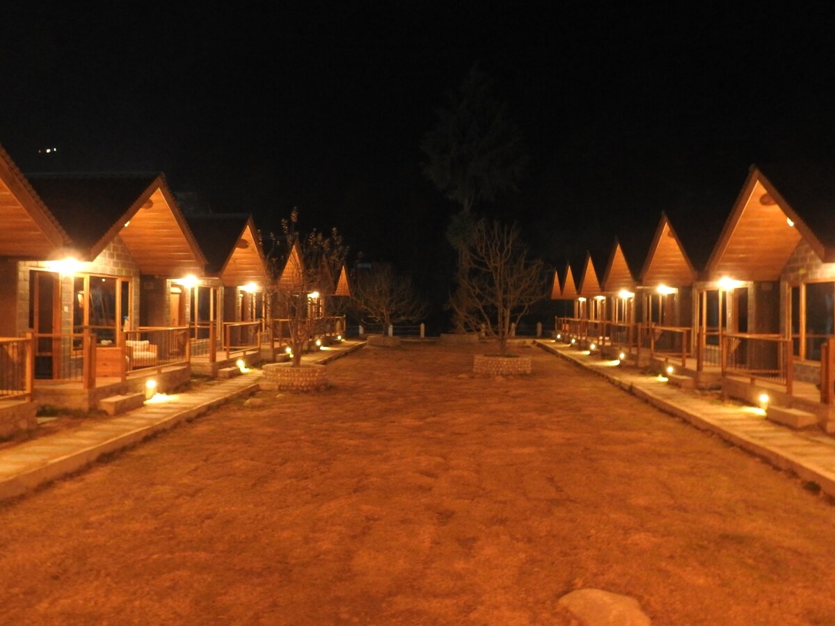 A row of vintage cottages is softly illuminated by warm lights at night, creating a welcoming ambiance. A well-maintained pathway leads to each cottage, bordered by a grassy area. Trees and landscaped elements add to the serene atmosphere of the resort.