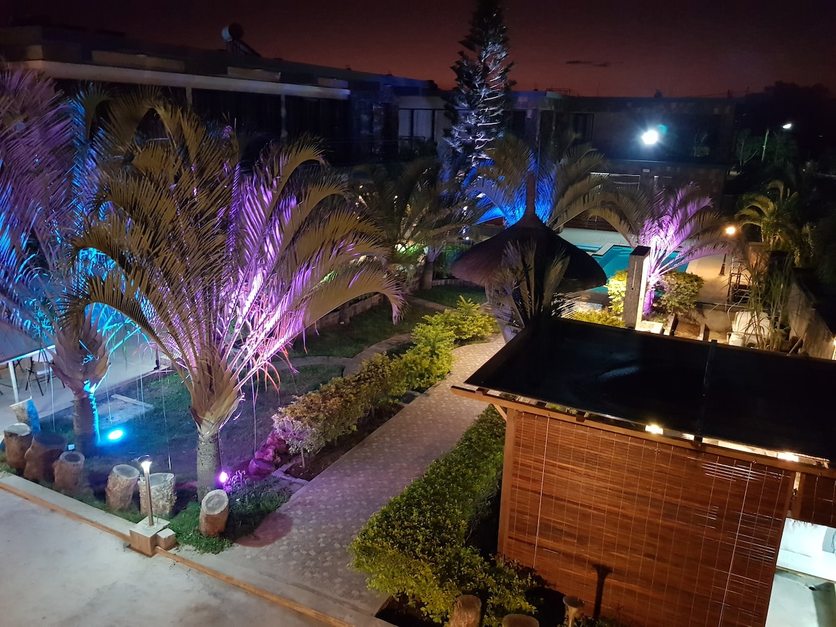 The outdoor space showcases a well-manicured garden area, featuring illuminated palm trees and a serene pathway. A sparkling swimming pool is visible amidst vibrant landscaping, accompanied by soft lighting that enhances the peaceful ambiance of the villa compound.
