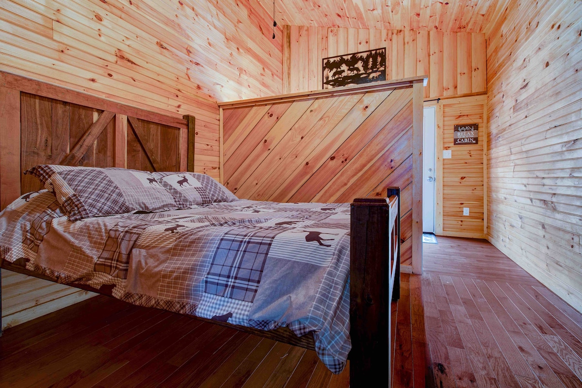 An open sleeping area is presented with a handmade full bed, featuring a patterned duvet cover. The walls are clad in warm wooden panels, with natural light illuminating the space. A door and additional decor can be seen in the background.