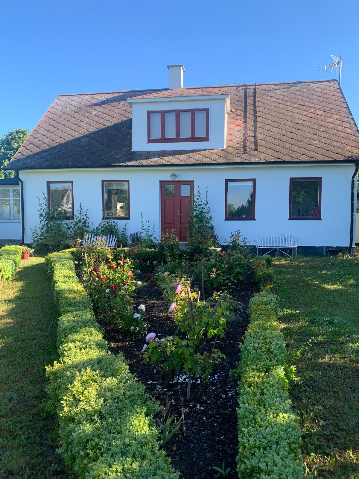 A traditional countryside home is featured, with a gabled roof and bright red accents around the windows and door. Well-maintained flower beds are bordered by trimmed hedges, showcasing a variety of blooming plants. The garden space surrounds the welcoming entrance of the house.