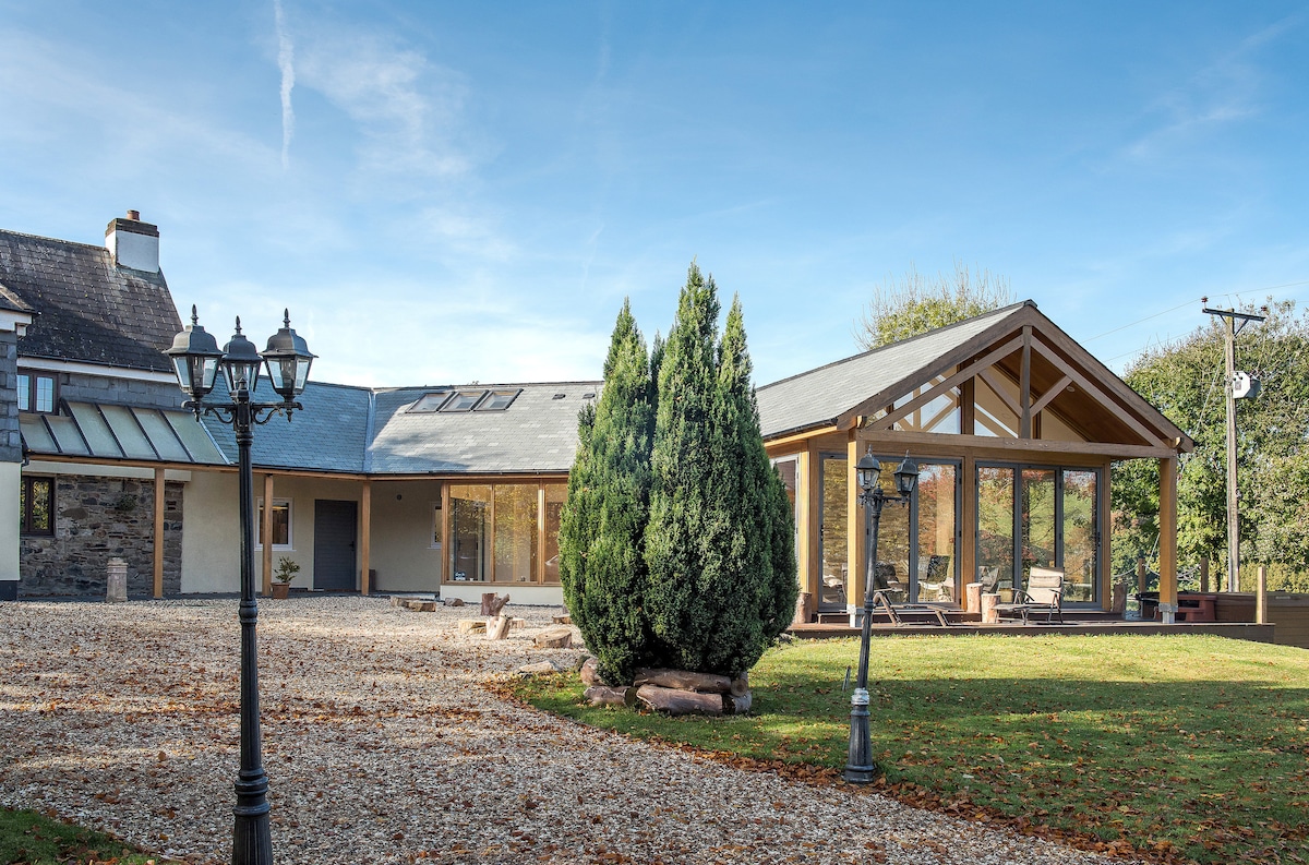 The exterior of the holiday cottage features a spacious patio area, framed by tall greenery, and complemented by ornamental lampposts. The building showcases a contemporary design with large glass sections, allowing for abundant natural light and a connection to the surrounding landscape.
