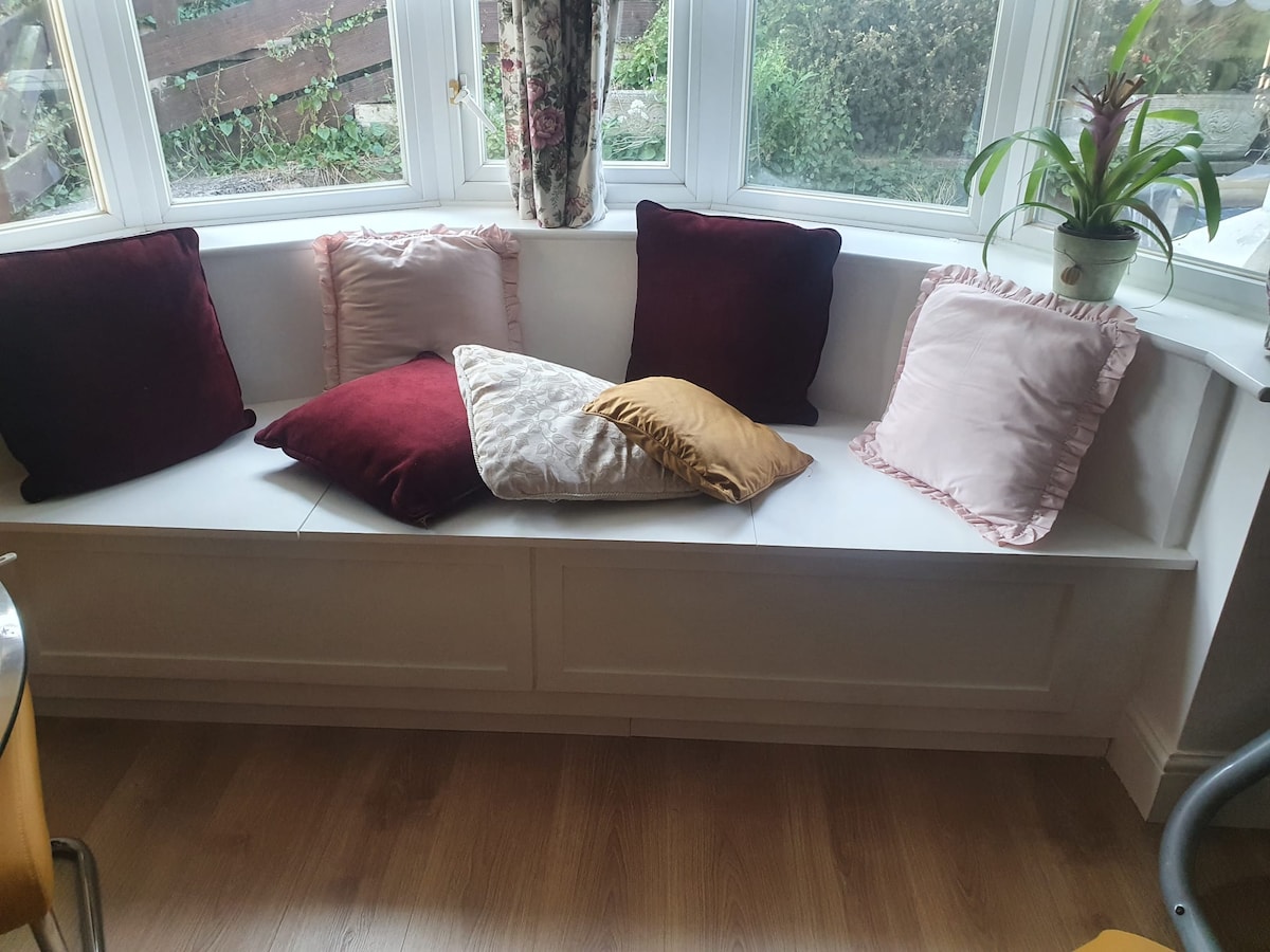 A comfortable window seat is fitted with an assortment of cushions in varying colors, including deep reds and soft pinks. Natural light spills in through the large windows, illuminating the cozy seating area. A potted plant adds a touch of greenery nearby.