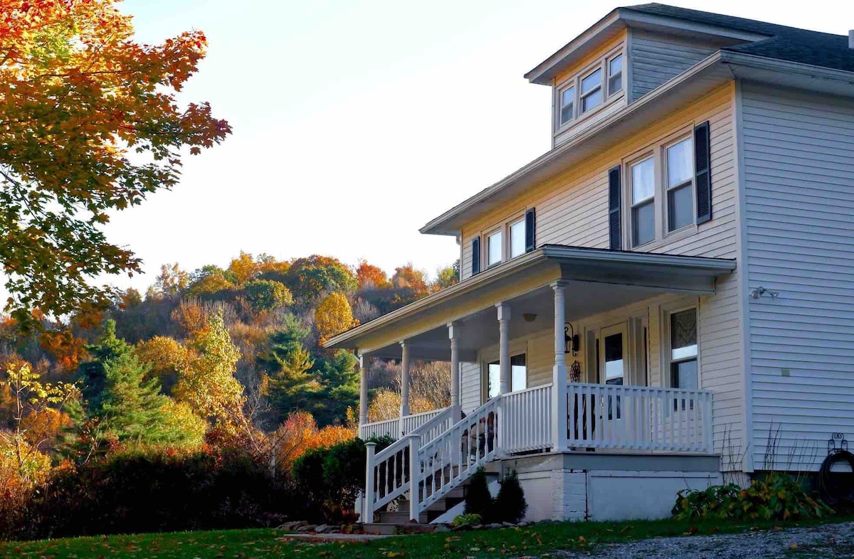 A charming two-story house is presented, featuring a covered front porch with white railings. Autumn foliage decorates the surrounding landscape, showcasing vibrant colors. The windows are framed in white, and the roofline adds a classic touch to the home's exterior.