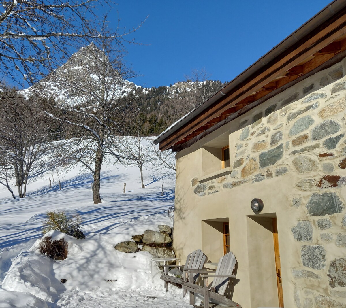 A rustic stone cottage is viewed against a clear blue sky, surrounded by snow-covered ground and leafless trees. A mountain peak rises in the background, completing the serene winter landscape. Simple outdoor seating is positioned at the cottage's entrance.