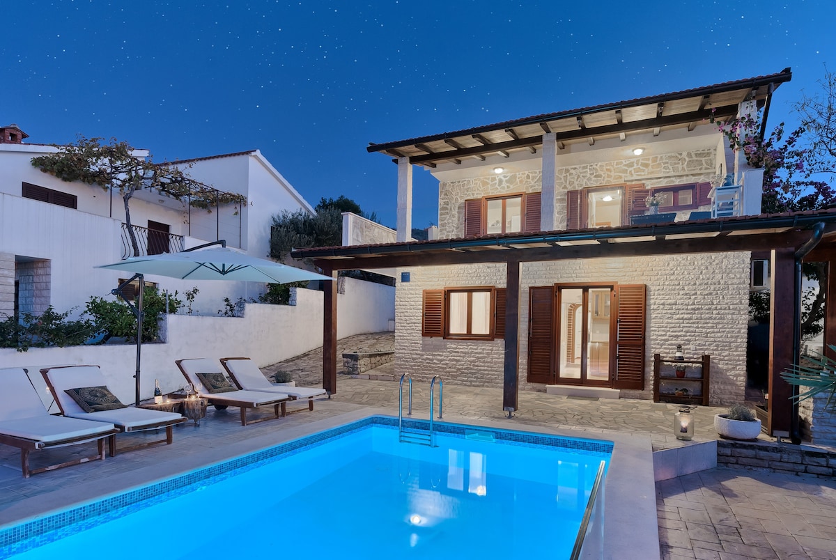 The two-story house is illuminated against a starlit sky, featuring a spacious outdoor area with a clear swimming pool. Wooden loungers are arranged near the poolside, providing a tranquil setting for relaxation. The architecture blends traditional elements with a modern touch.