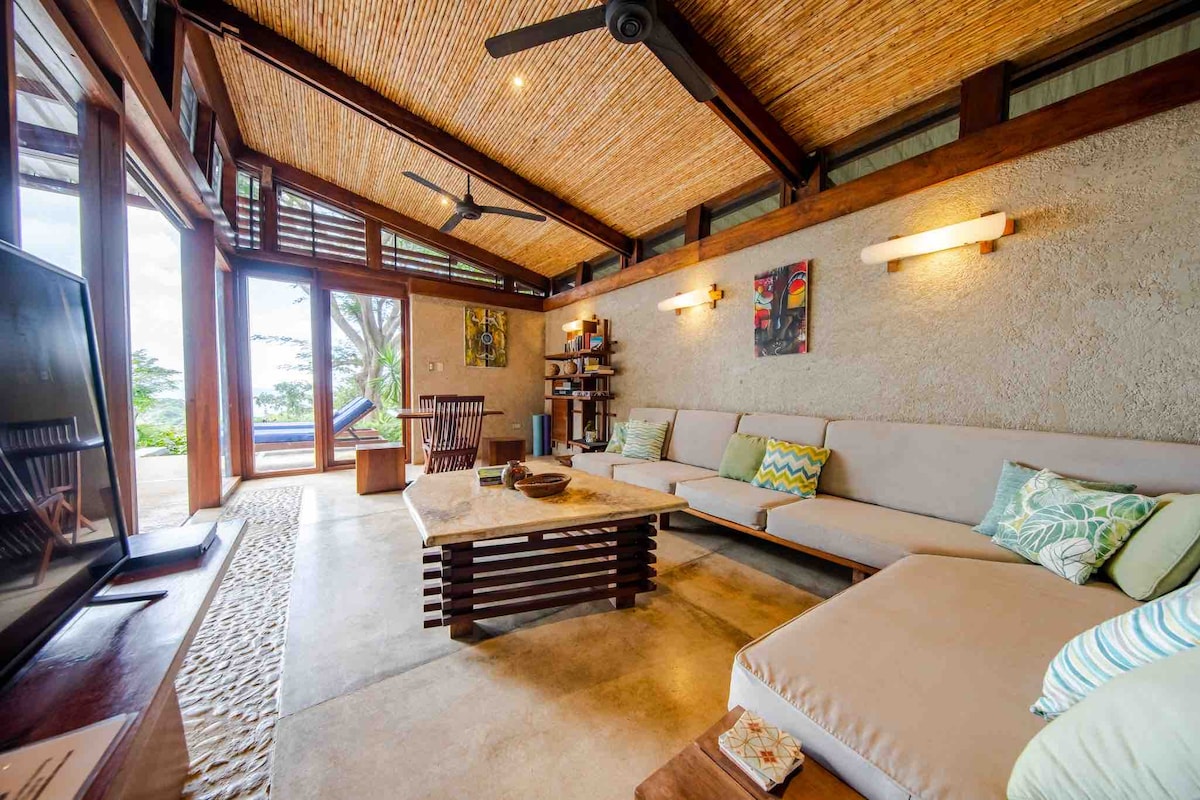 A spacious living area features a comfortable sectional sofa paired with decorative cushions. Natural light enters through large glass doors, offering views of the outdoors. A stone coffee table sits in the center, surrounded by a wooden dining table and simple wall art.
