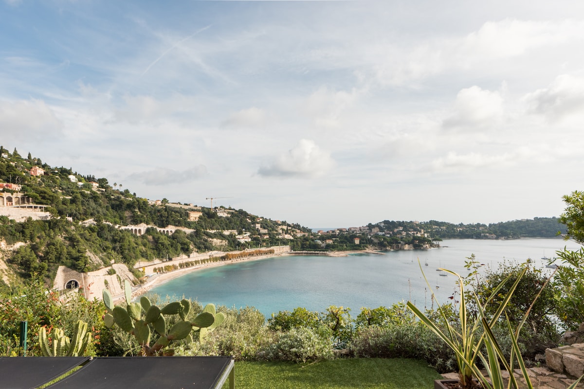A serene view of the coastline is captured, showcasing gentle waves lapping against the shore. Lush greenery and a garden are visible in the foreground, complementing the expansive vista of the calm sea and distant hills.