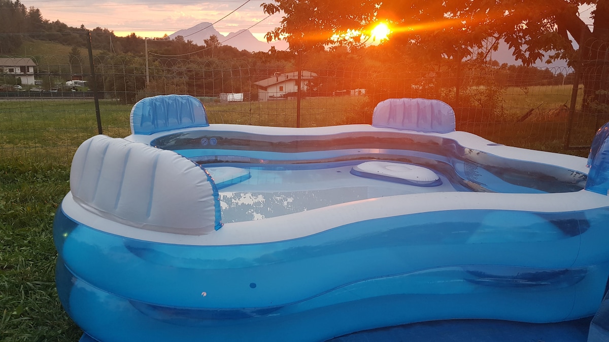 An inflatable pool is depicted, filled with clear water and surrounded by green grass. The sun sets in the background, casting an orange glow, while the distant mountains frame the scene. Comfortable seating areas are integrated into the pool's design.