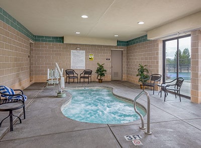 An indoor hot tub is surrounded by several chairs arranged nearby. Large windows allow natural light to fill the area, and potted plants add a touch of greenery to the space. Safety signage is visibly posted on the wall.