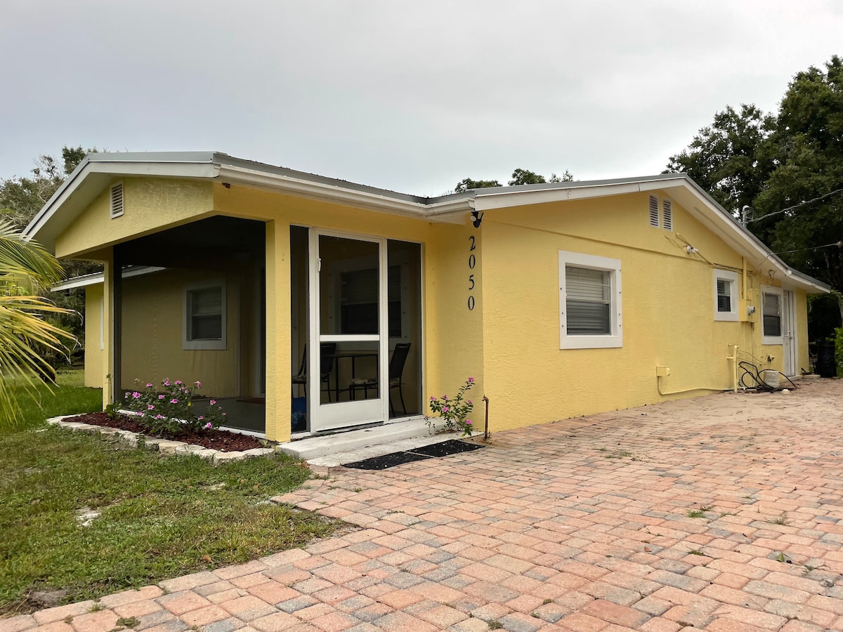 A bright yellow exterior of a single-story home is shown, featuring an inviting covered porch with a screened area. The driveway is made of pavers, bordered by a small garden with flowers. Surrounding greenery complements the setting.