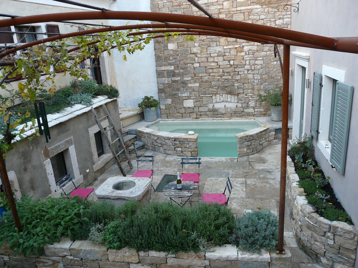 A courtyard is presented, featuring a stone plunge pool surrounded by a small paved area. Seating arrangements with black metal chairs and pink cushions are visible, along with a stone fire pit. Lush greenery and herbs flank the edges, complemented by the rustic stone walls of the setting.