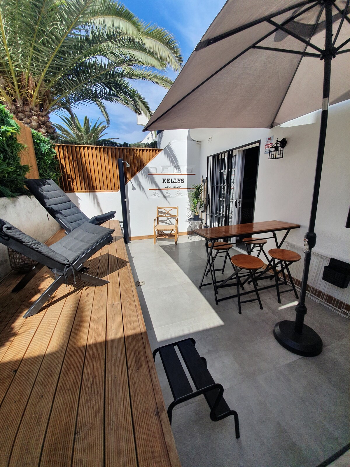 An outdoor terrace is presented, featuring a wooden deck and comfortable seating arrangements. A dining table with chairs is positioned under an umbrella, alongside a reclining lounge chair for relaxation. Lush palm trees provide a tropical backdrop, enhancing the serene atmosphere.