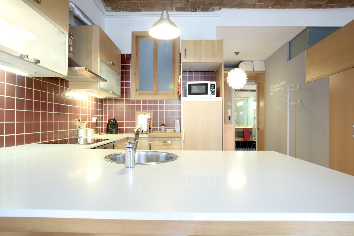 A well-equipped kitchen area features a modern countertop with a sink, complemented by wooden cabinetry and a vibrant patterned backsplash. Appliances such as a microwave and kettle are present, creating a functional space. A seamless flow connects to the living area in the background.