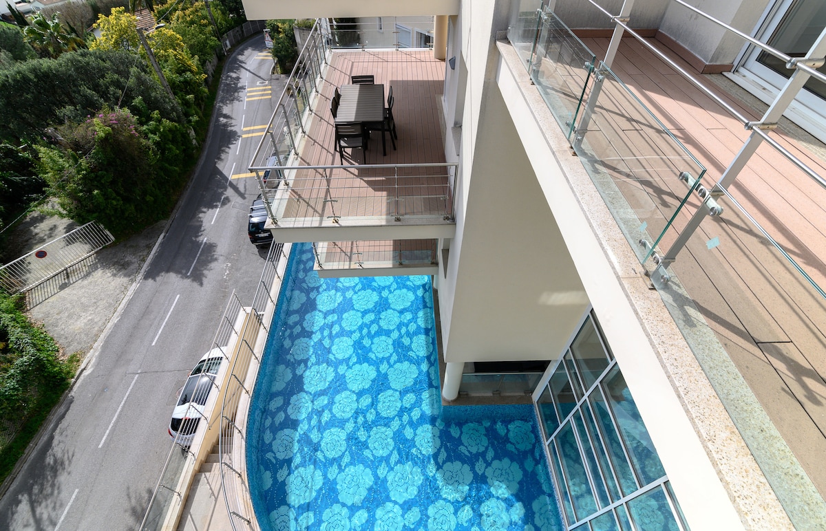 A spacious terrace is highlighted, featuring a table and chairs. Below, a shared pool showcases an intricate blue patterned surface. The surrounding greenery and pathway are visible, offering a glimpse of the residential area.