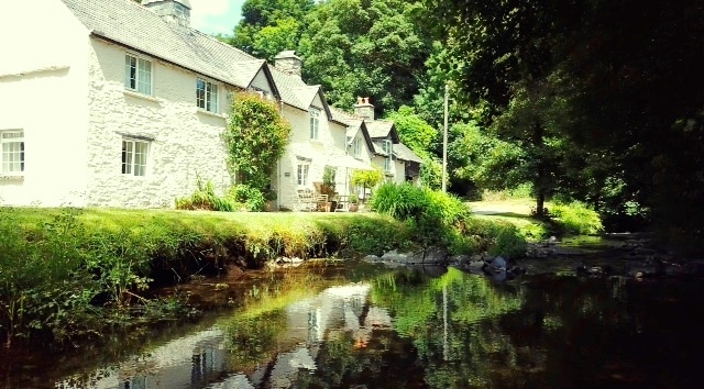 A collection of charming white cottages is situated alongside a tranquil stream, reflecting the surrounding greenery. Lush grass and trees frame the scene, offering a peaceful ambiance. Outdoor seating can be seen on the cottage porch, inviting relaxation in the natural setting.