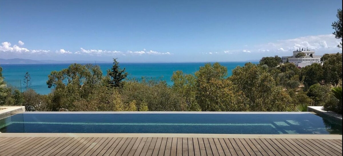 An infinity pool stretches across the foreground, blending seamlessly into the distant turquoise waters of the bay. Lush greenery frames the scene, with trees dotting the landscape. A clear sky with soft clouds enhances the expansive view over the coastline.