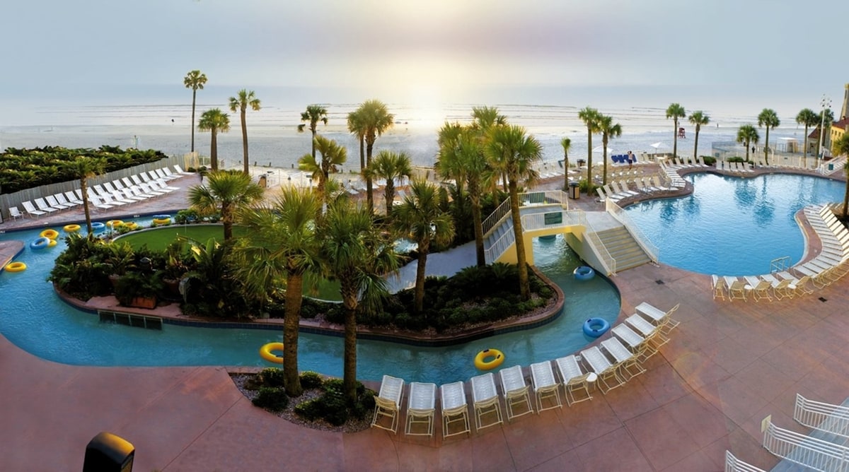 A tranquil pool area is framed by lush palm trees and features a lazy river winding through the resort. Lounge chairs line the poolside, while colorful flotation rings rest in the water. The sunset creates a gentle glow over the ocean in the background.