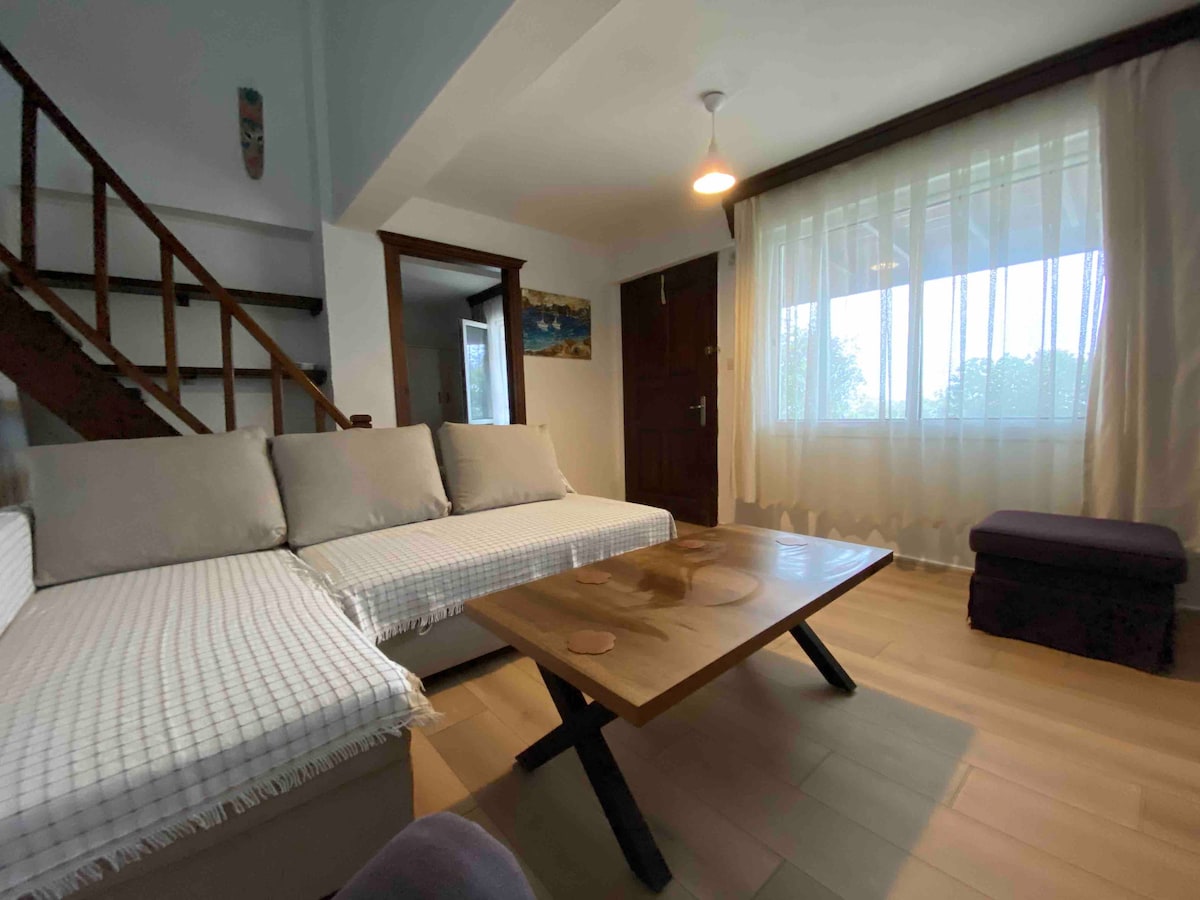 A bright living area is presented with a comfortable, L-shaped sofa adorned with light-colored cushions. A wooden coffee table sits in front, complemented by natural light streaming through a large window draped with sheer curtains. A staircase is visible in the background.