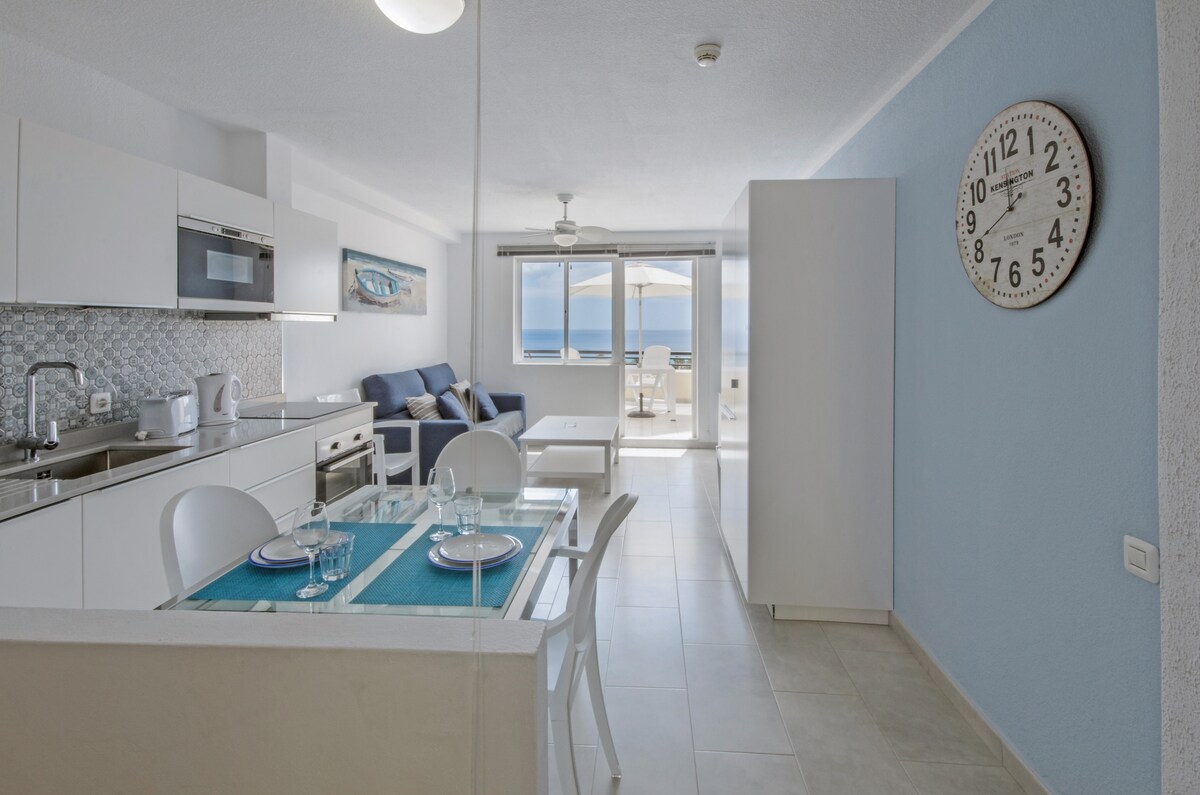 An open-plan living space features a modern kitchen equipped with stainless steel appliances and a dining area with a glass table set for two. A comfortable seating area is positioned against a light blue wall, offering views through large windows that lead to a terrace.