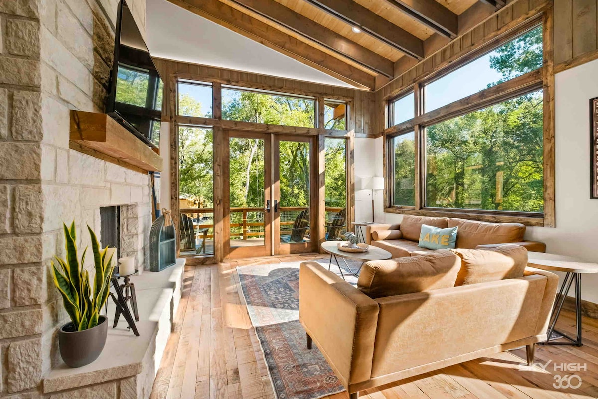 An open living area is presented with vaulted wooden ceilings and large windows that frame views of the outdoors. A comfortable sofa is arranged with a soft area rug underneath. A flat-screen TV is mounted on the wall above a stone fireplace, creating a relaxed atmosphere.