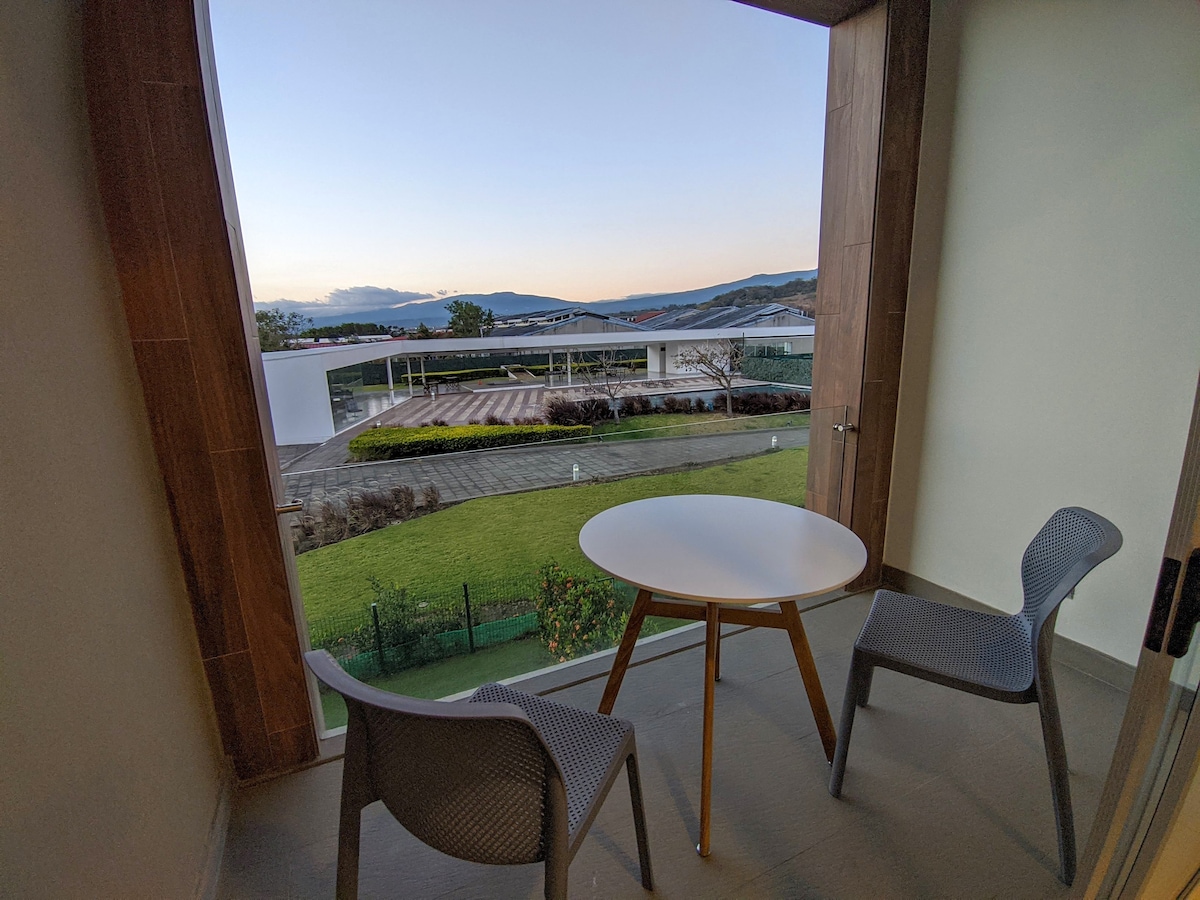 A small balcony features a round table accompanied by two chairs, offering a view of landscaped green spaces and distant mountains. Natural light enters through large windows, enhancing the sense of openness in the area.