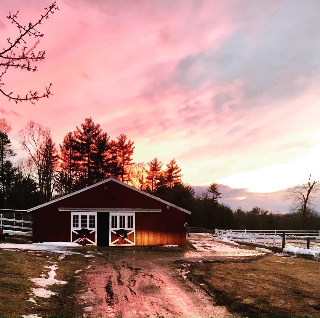 A red barn stands against a dramatic sunset, with hues of pink and orange coloring the sky. Snow remnants are visible on the ground, leading up to the barn entrance. Trees frame the background, creating a serene rural landscape.