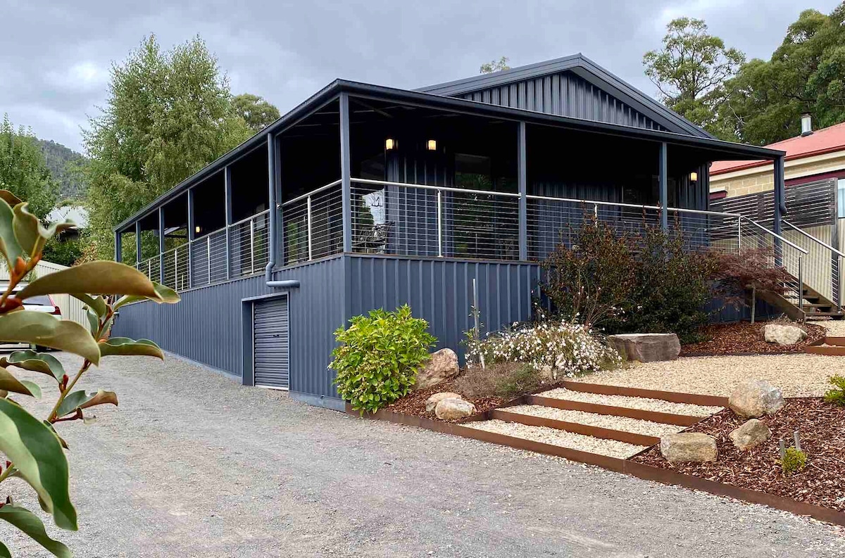 The exterior of a blue-clad house is shown, featuring a spacious deck with railings. Surrounding greenery and landscaped rocks are visible, enhancing the outdoor setting. The stone pathway leads to the entrance, complemented by various shrubs and plants for an inviting appearance.