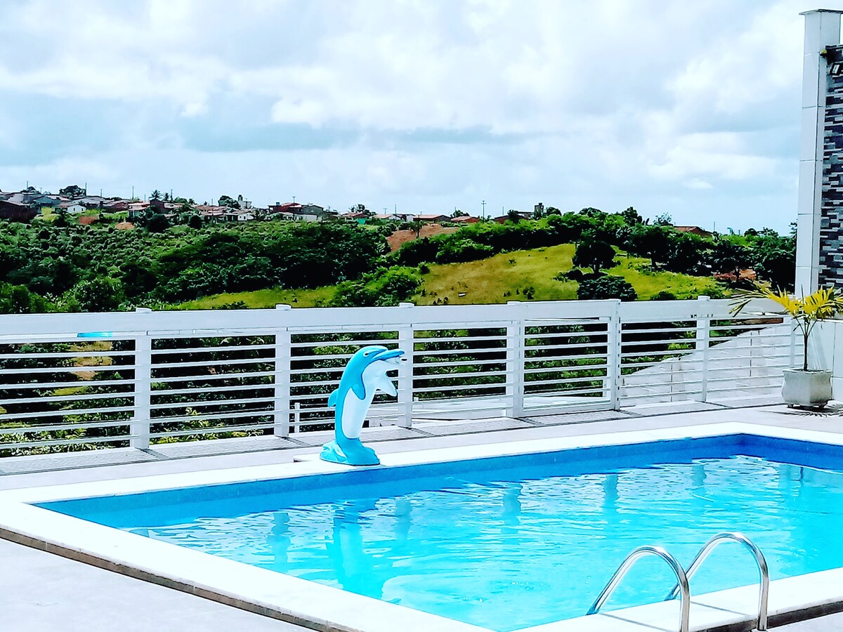 A swimming pool with clear blue water is bordered by a spacious deck. A dolphin-shaped float is positioned in the water, while a scenic view of lush green hills is visible in the background, separated by a low white railing.