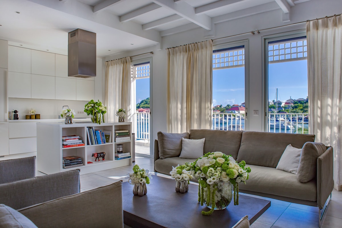 A bright living area is presented, featuring large windows with light flowing in from outside. Soft drapes frame the windows, while a neutral-toned sofa is complemented by a simple wooden coffee table adorned with fresh flowers. A bookshelf holds an array of books, enhancing the cozy ambiance.
