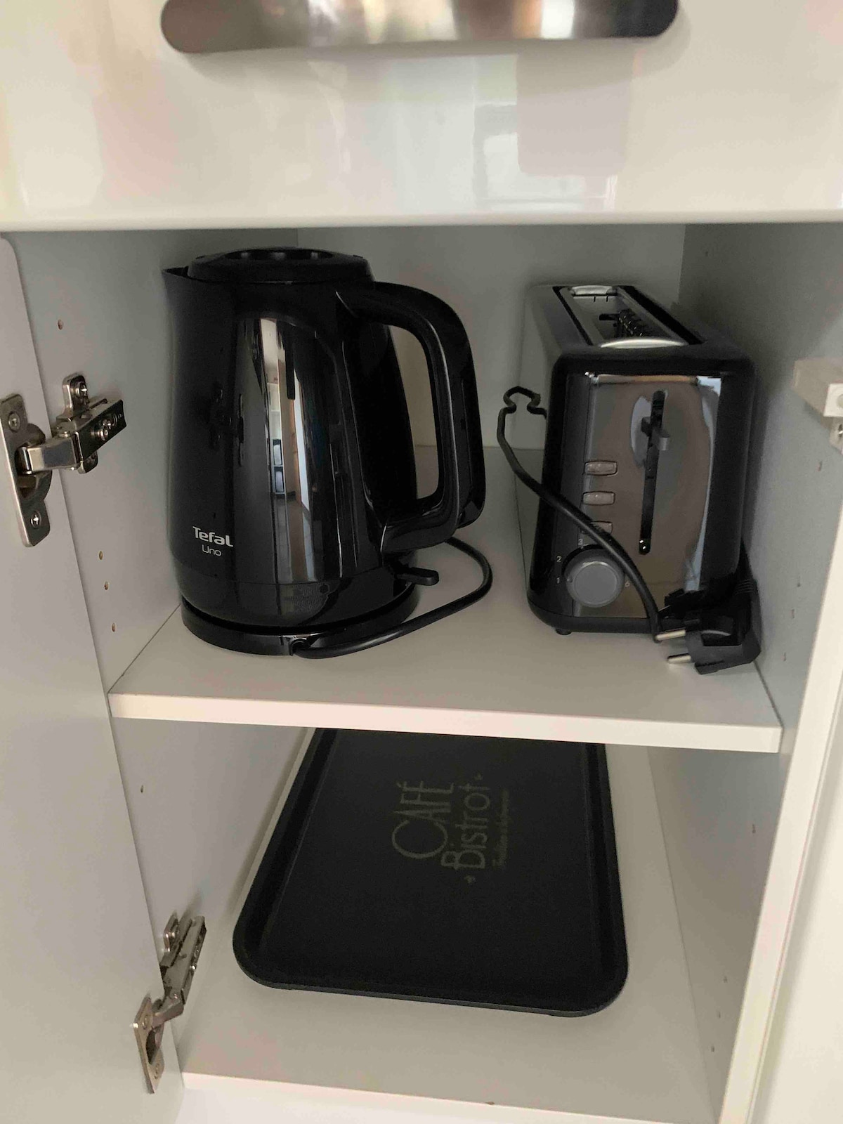 A kitchen cabinet is shown with a black kettle and a silver toaster on the shelf. A tray with the text 'Café Bistrôt' is located on the bottom shelf. The cabinet doors are closed but display a clean, modern interior.