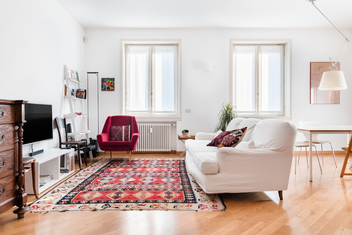 A bright living room features a mix of modern and antique furnishings. A large white sofa and a red accent chair are situated on a patterned rug. Shelves hold various books and decor, while a TV is positioned against a light-colored wall, complemented by two large windows.