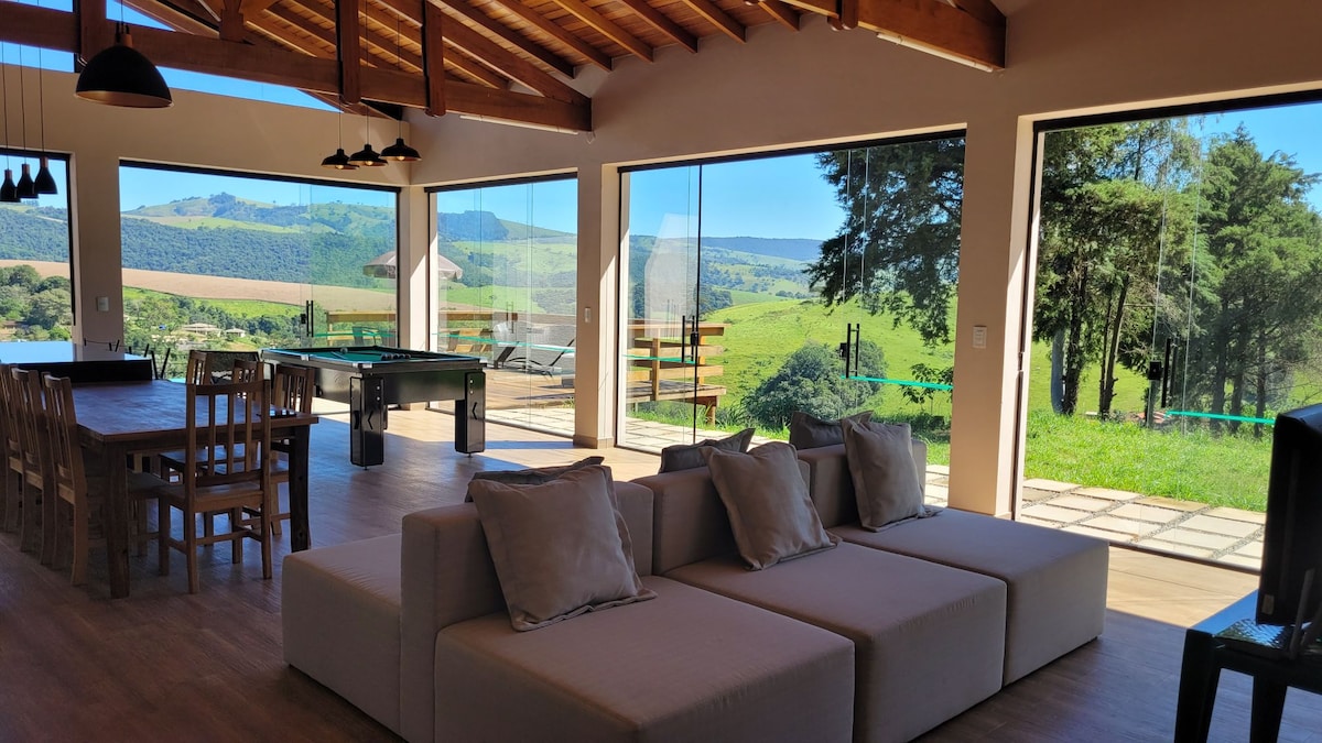 A spacious interior is highlighted by large glass windows, allowing natural light to fill the room and providing a view of the surrounding green landscape. A comfortable seating area features plush cushions, and dining furniture is arranged nearby, creating an inviting space for relaxation and gatherings.